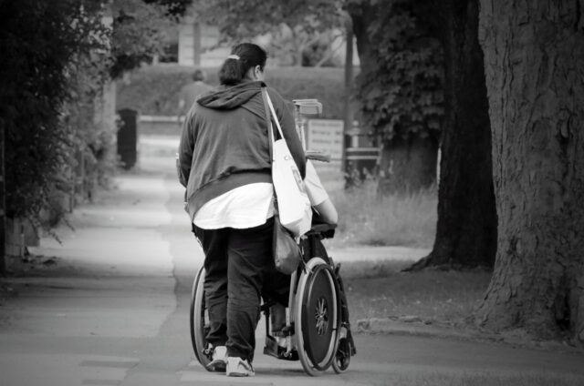 Grafika przedstawia osobę pchającą wózek inwalidzki przez alejki parkowe.