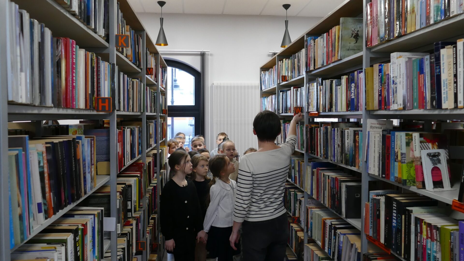 Dzieci z panią bibliotekarką oglądające książki na półkach bibliotecznych.