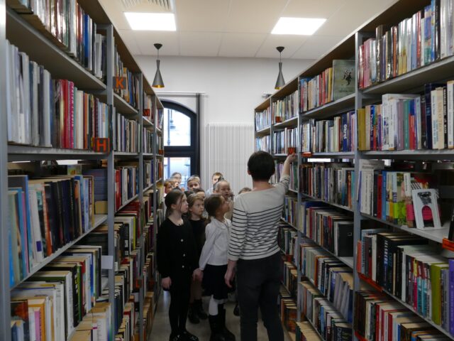 Dzieci z panią bibliotekarką oglądające książki na półkach bibliotecznych.