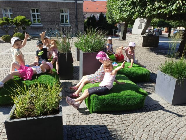 Strefa Relaksu na pleszewskim Rynku składająca się z poduszek imitujących trawę i donic z roślinnością oraz siedzące na niej dzieci.